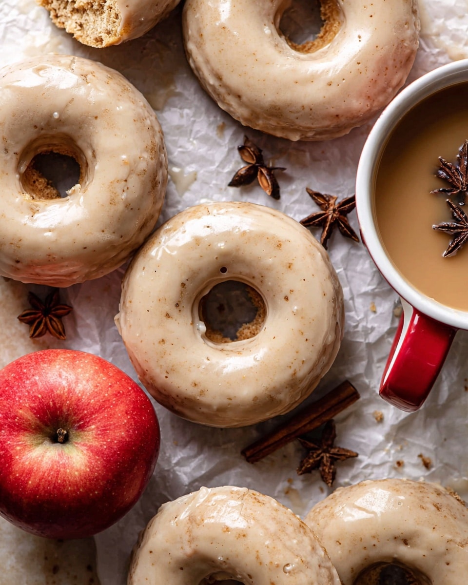 A close-up view of several glazed donuts with a smooth light brown icing layer covering each ring-shaped donut, showing a slightly soft and crumbly texture inside from broken pieces. The glaze has a matte and creamy finish with tiny specks, topped neatly and evenly. A bright red apple with a shiny skin rests nearby, adding a fresh contrast. The donuts are placed on crumpled white parchment paper over a white marbled surface. To the side, a white cup with a red handle holds a light brown drink, with star anise floating on the surface and scattered around for decoration. Photo taken with an iphone --ar 4:5 --v 7
