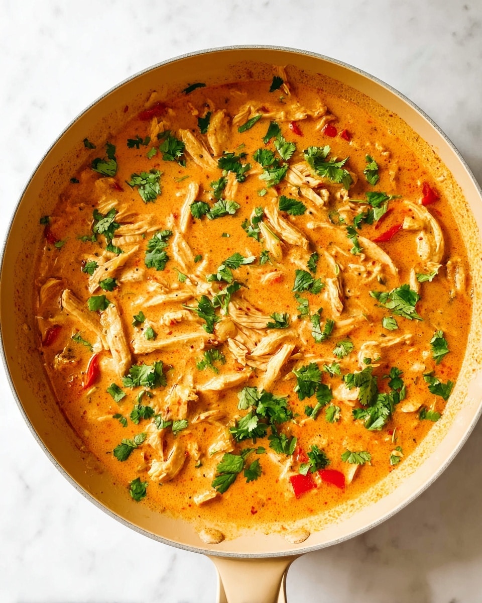 A large pan filled with a creamy orange curry sauce covering tender strips of cooked chicken and small pieces of red bell pepper, all topped with bright green chopped cilantro leaves scattered evenly across the surface. The sauce has a smooth texture with hints of creaminess and slight speckling from spices. The pan has a light beige handle and sits on a white marbled surface, giving a clean, bright background to the rich colors of the curry. photo taken with an iphone --ar 4:5 --v 7