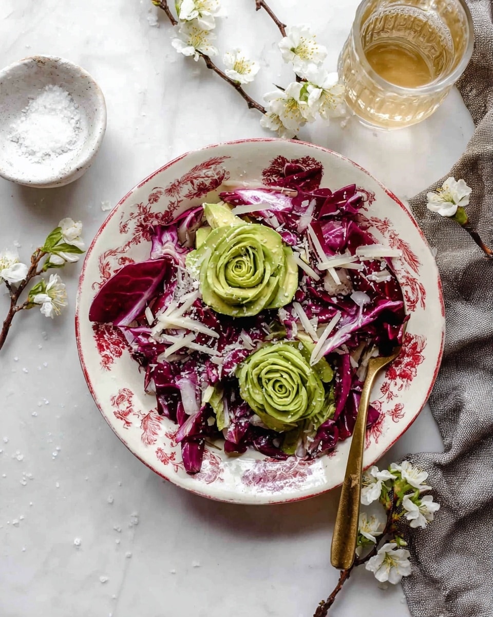 A white plate with a red floral pattern holds a salad with two rose-shaped green avocado slices placed on top of a base of deep purple radicchio leaves. Shredded white cheese is sprinkled over the radicchio in uneven pieces, adding a contrast in color and texture. A gold spoon rests on the edge of the plate. The plate is set on a white marbled surface with small sprigs of white blooming flowers placed around it, along with a small white bowl of salt, a glass with light amber liquid, and a gray cloth. Photo taken with an iphone --ar 4:5 --v 7