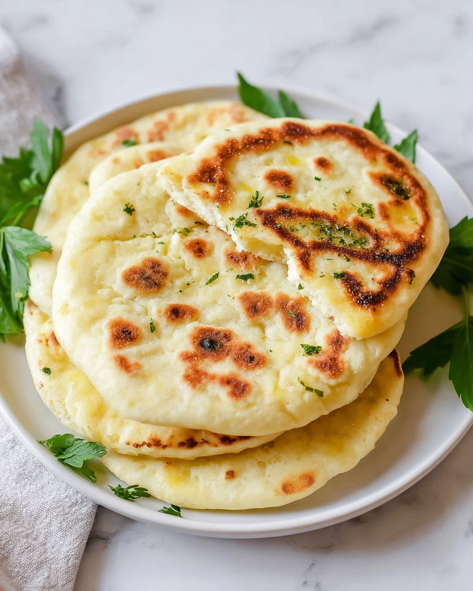 A white plate holds a stack of four pieces of flatbread with a golden-brown toasted pattern on the surface. The top flatbread is slightly torn to show its soft, fluffy inside with small green herb bits sprinkled on it. The flatbreads are thick and have a light cream-yellow color with darker browned spots from cooking. The plate rests on a white marbled surface and is garnished with fresh green leaves around the edges. Photo taken with an iphone --ar 4:5 --v 7