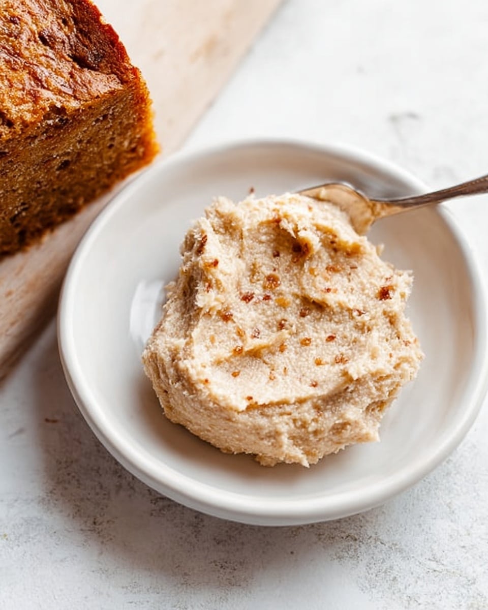 A small white plate holds a scoop of creamy, light beige almond butter with visible small bits of almond throughout, giving it a slightly rough texture. On the right side of the image, a golden-brown loaf of bread with a crunchy crust is partially visible. The whole scene rests on a white marbled surface with faint gray veins. Photo taken with an iphone --ar 4:5 --v 7