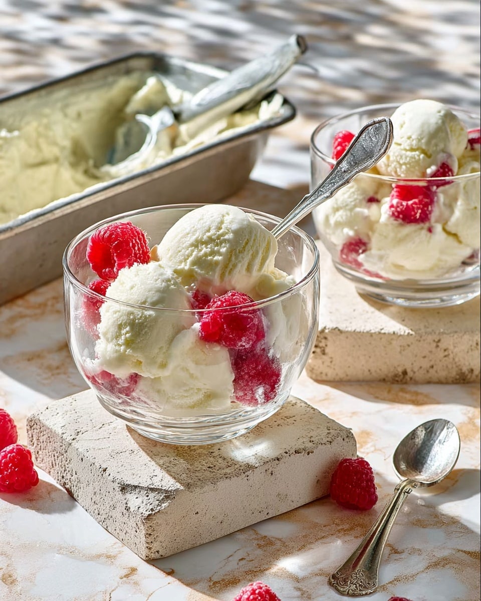 Two clear glass dessert cups each contain about two scoops of creamy white ice cream, with bright red raspberries nestled around the ice cream inside the cups. One cup is in the front and has a silver spoon sticking out of the ice cream. The cups rest on rough, light stone square bases. In the background, a silver container holds more ice cream, and a silver spoon with some ice cream lies nearby on a white marbled surface. The scene is lit brightly, causing soft shadows and a warm, inviting look. photo taken with an iphone --ar 4:5 --v 7