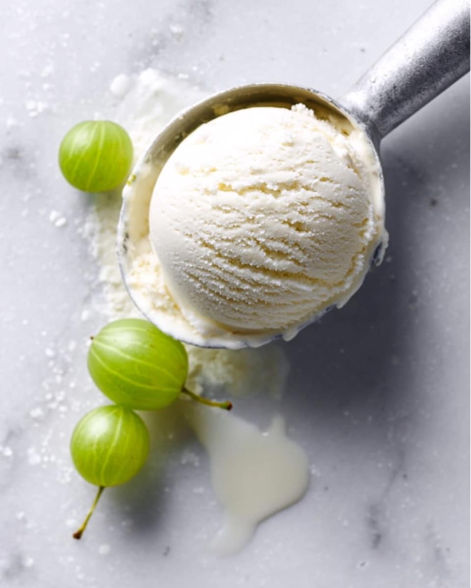 A single scoop of creamy white ice cream with a smooth and slightly cracked texture sits in a silver ice cream scoop resting on a white marbled surface. Next to the scoop, there are two small green gooseberries with a shiny texture, giving a fresh look to the scene. A small drip of melted ice cream runs down from the scoop onto the marbled surface, adding a natural touch. The photo taken with an iphone --ar 4:5 --v 7