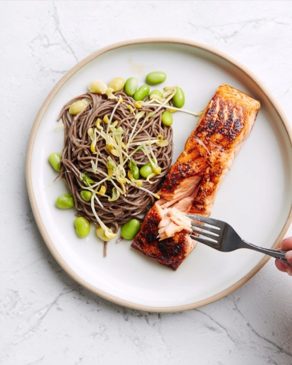 A white plate on a white marbled surface holds two main parts: on the left, a nest of brown soba noodles mixed with green edamame beans and light green baby corn, creating a fresh and slightly shiny texture; on the right, a thick, grilled salmon fillet with a golden-brown crust and juicy inner pink layer, where a woman’s hand holds a fork cutting a small piece from the salmon, showing the flaked fish texture inside. Photo taken with an iphone --ar 4:5 --v 7