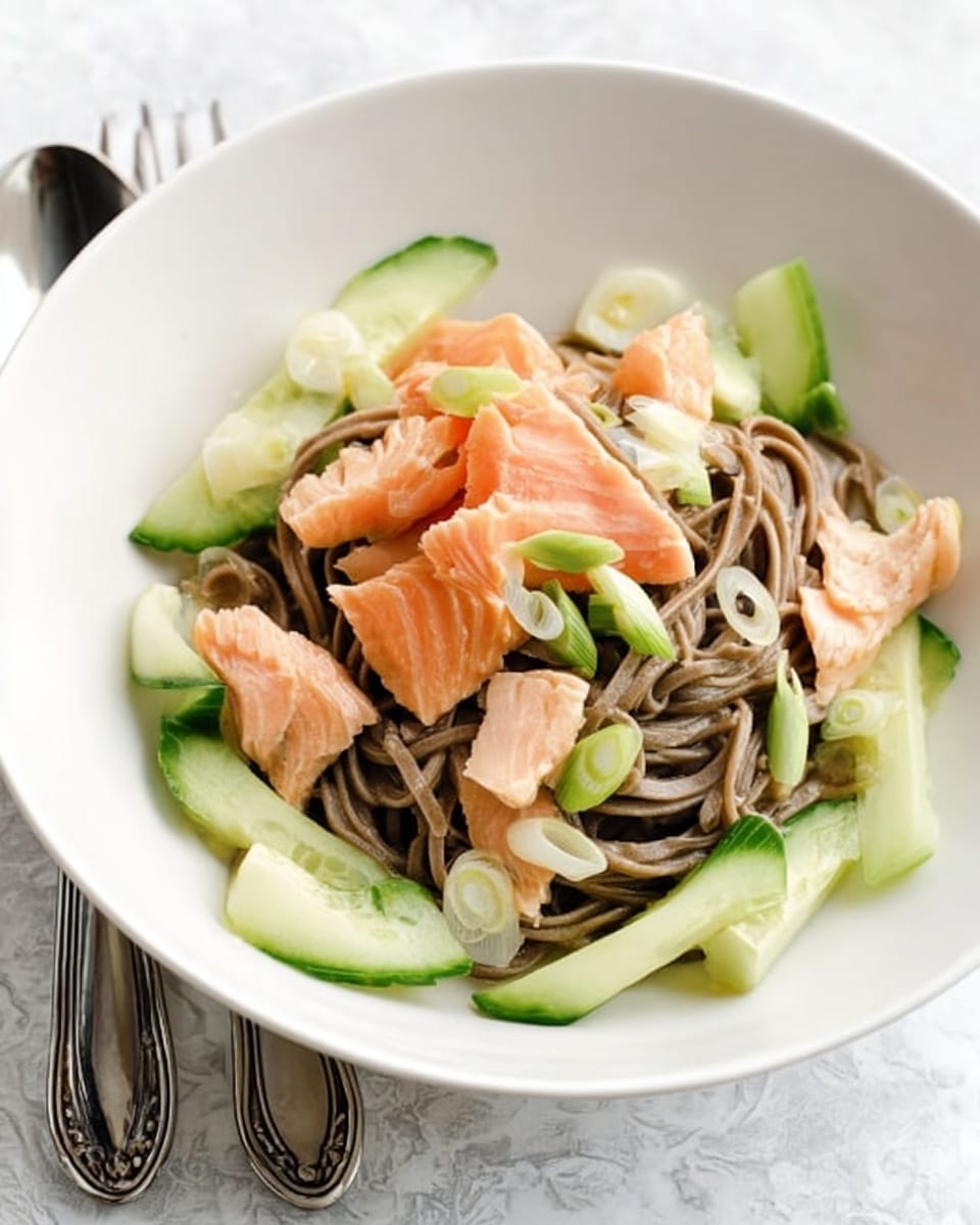 The image shows a white bowl with three main layers: the bottom layer has dark brown soba noodles with a smooth texture, the middle layer has sliced light green cucumbers placed around the noodles, and the top layer has pieces of light pink cooked salmon along with sliced light green scallions scattered evenly. The bowl is placed on a white marbled texture surface with two silver utensils resting beside it. photo taken with an iphone --ar 4:5 --v 7