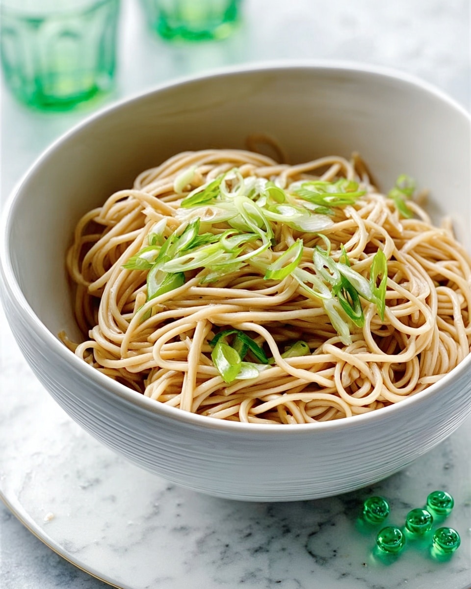 A white bowl filled with a single layer of light brown noodles, topped with thin, bright green sliced scallions, sitting on a white marbled surface with a few green glass beads next to the bowl. The noodles look smooth and slightly glossy, and the scallions are fresh and crisp, adding a sharp contrast in color and texture. The bowl has a simple, modern design with soft curves. photo taken with an iphone --ar 4:5 --v 7