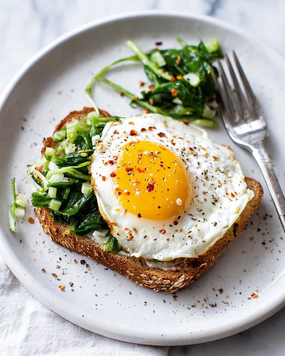 A toasted brown bread slice on a white plate forms the base layer, topped with a bright, sunny-side-up egg whose white edges are slightly wavy and golden around the bottom right corner; the egg yolk is vibrant yellow with small pepper flakes sprinkled on top. On one side of the egg, there is a layer of chopped, cooked greens in varying shades of green, adding texture and color contrast. The plate rests on a white marbled surface with some scattered pepper specks and a fork placed near the top of the plate holding a small portion of greens. photo taken with an iphone --ar 4:5 --v 7