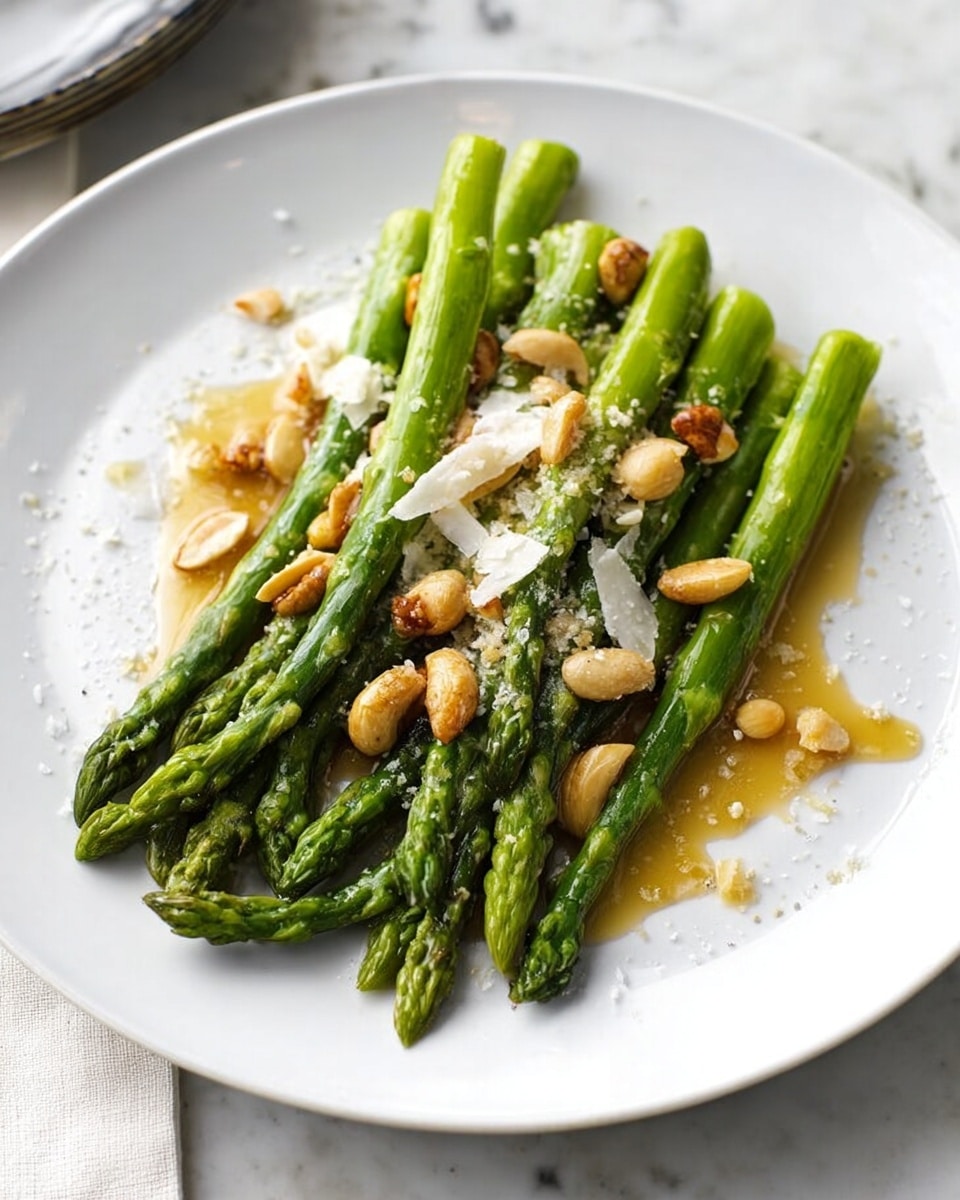 A simple dish shows a neat pile of bright green asparagus spears laid across the center of a white plate placed on a white marbled surface. The asparagus is topped with a light drizzle of golden brown sauce and sprinkled with off-white shaved cheese and a few scattered roasted or toasted nuts that add texture and contrast. The overall look is fresh and light with natural colors and soft lighting that make the asparagus appear glossy and tender. Photo taken with an iphone --ar 4:5 --v 7