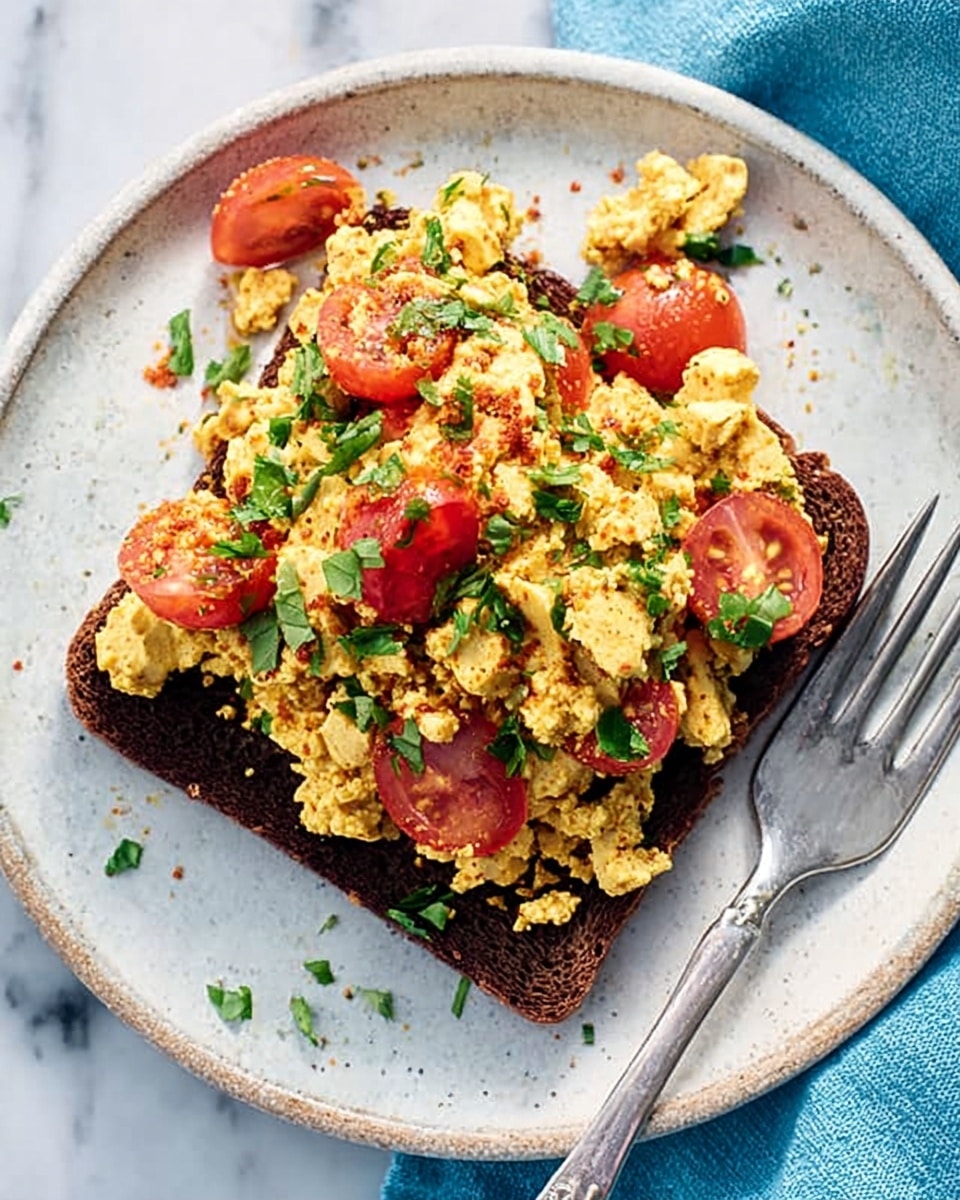 The image shows a single slice of dark brown bread topped with a colorful scrambled tofu mix. The scrambled tofu is light yellow with a crumbly texture and is mixed with bright red cherry tomato halves and bits of green herbs scattered on top. The dish is served on a white plate with a subtle rustic texture. A silver fork rests on the right side of the plate, which is placed on a white marbled surface with a blue cloth partially visible in the top right corner. Photo taken with an iphone --ar 4:5 --v 7
