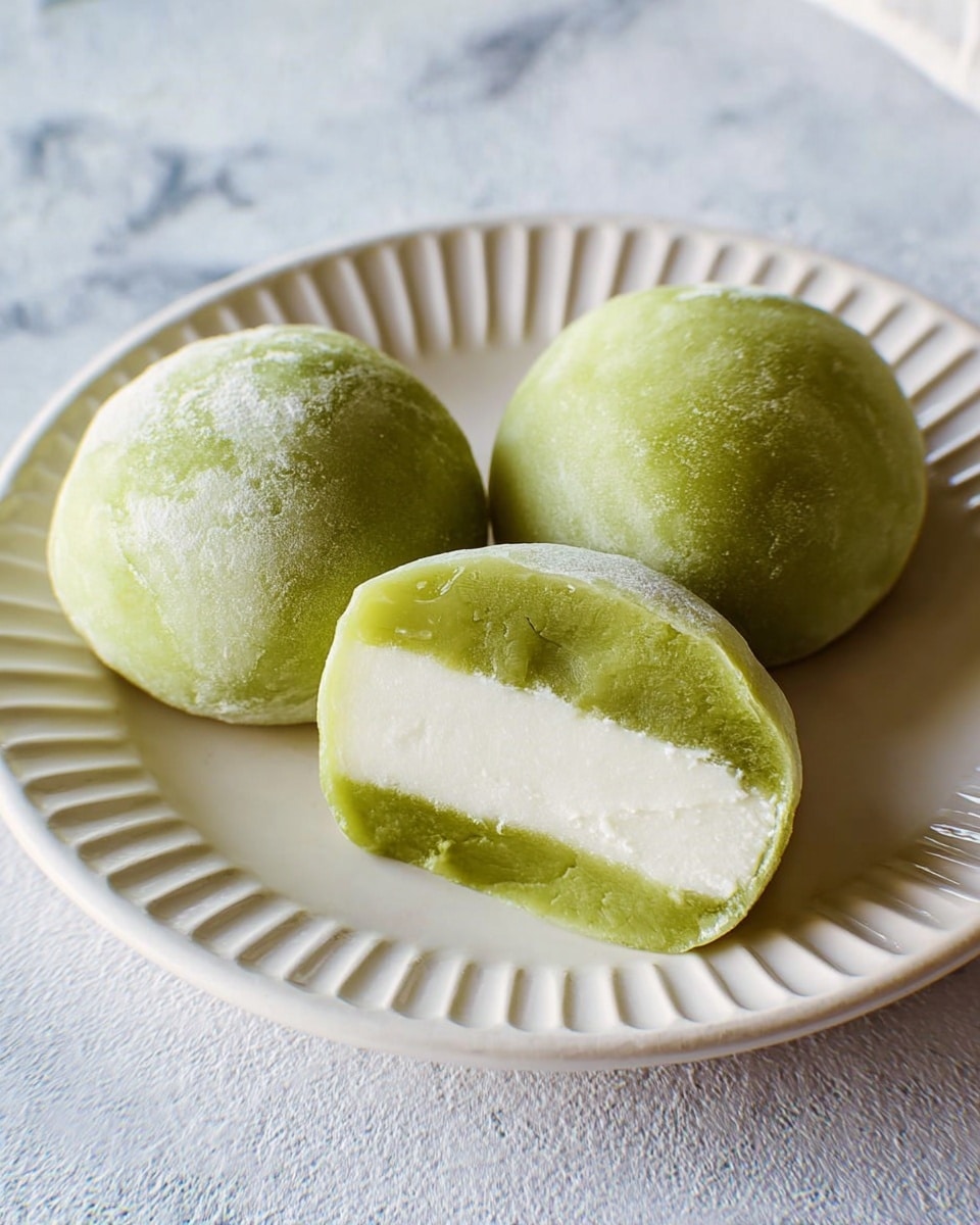 Three green mochi balls sit on a white plate with a ridged edge, placed on a white marbled textured surface. Two whole mochi balls have a smooth, slightly powdery green outer layer with a soft texture. One mochi is cut in half, showing two layers inside: a thick, dense white creamy center, and a thinner smooth green layer encasing it. The overall look is soft and fresh. photo taken with an iphone --ar 4:5 --v 7