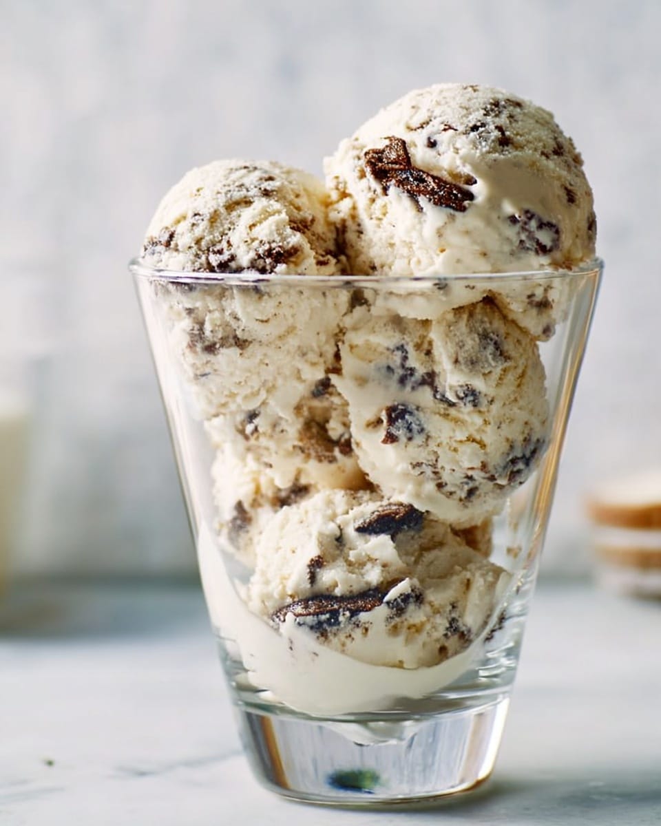 A clear glass filled with four large scoops of creamy ice cream that is light beige in color with dark brown chunks mixed throughout. The ice cream is layered from the bottom to the top of the glass, showing a creamy texture with small melted parts blending with the chunks. The background is white marbled texture, giving a clean and fresh look. Photo taken with an iphone --ar 4:5 --v 7