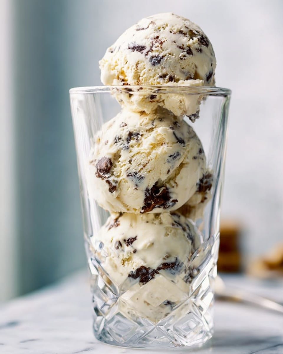 A clear, tall glass holds four round scoops of ice cream stacked unevenly. The ice cream is creamy white with dark brown chunks mixed throughout, showing a rich texture with bits of chocolate or fruit. The glass itself is faceted and catch the light, making the ice cream inside look shiny and inviting. The background is out of focus with soft light and a white marbled surface beneath the glass. photo taken with an iphone --ar 4:5 --v 7