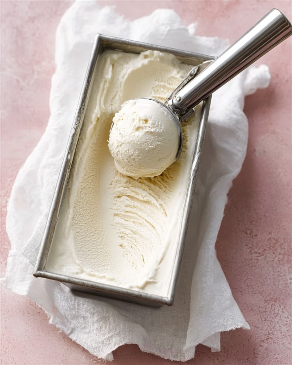 A shiny metal rectangular container filled with smooth, creamy white ice cream, with one round scoop being lifted by a metal ice cream scoop that has a long handle. The container rests on a white cloth napkin, all placed on a white marbled textured surface with a light pink background. The ice cream has a soft texture with gentle swirls on the surface. Photo taken with an iphone --ar 4:5 --v 7