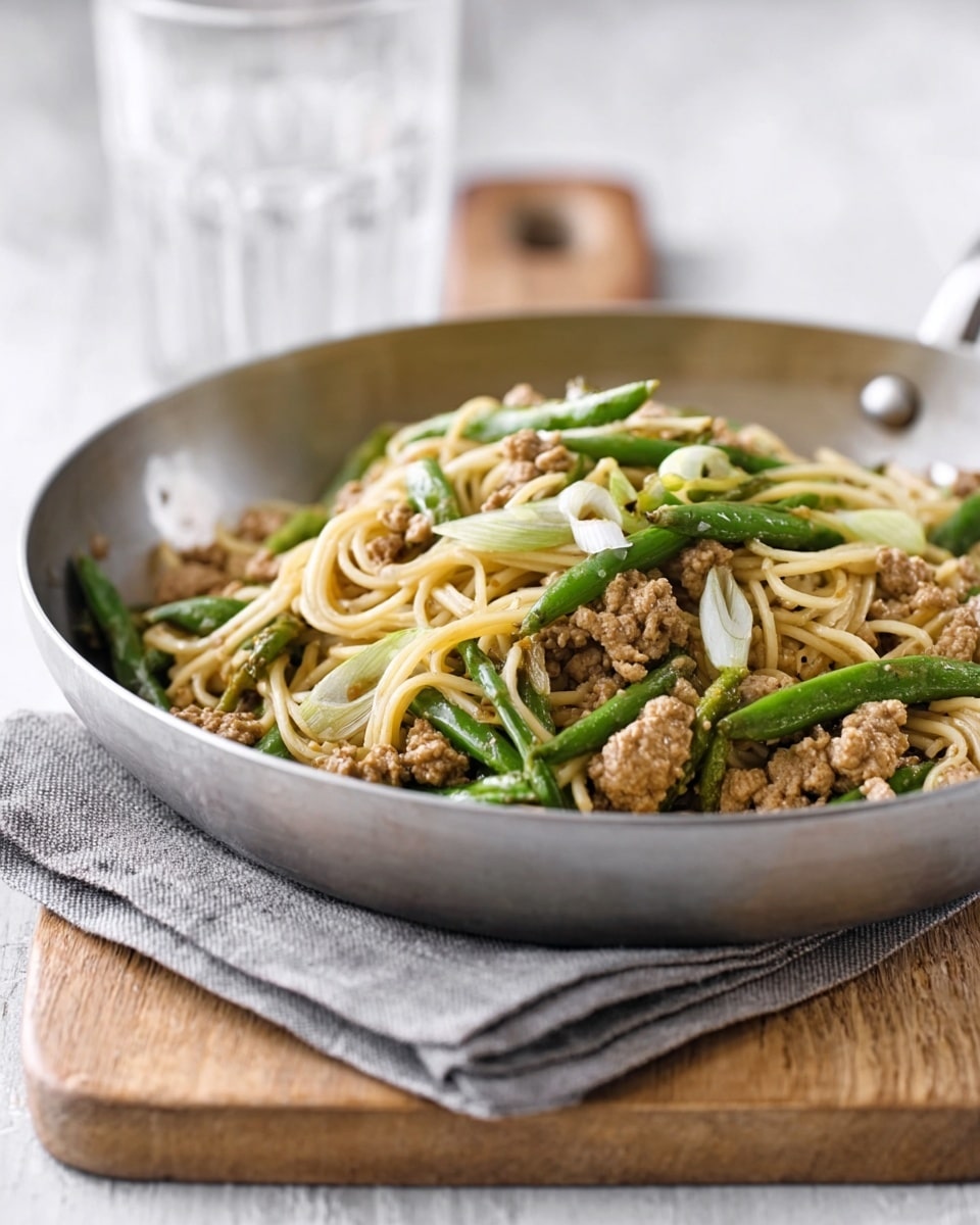 A metal pan filled with a dish of light brown cooked ground meat, mixed with light green chopped beans and white sliced spring onions on top of thin, pale yellow noodles. The pan is placed on a folded gray cloth napkin, which rests on a wooden board, all set on a white marbled surface. In the background, there is a blurry glass container. photo taken with an iphone --ar 4:5 --v 7