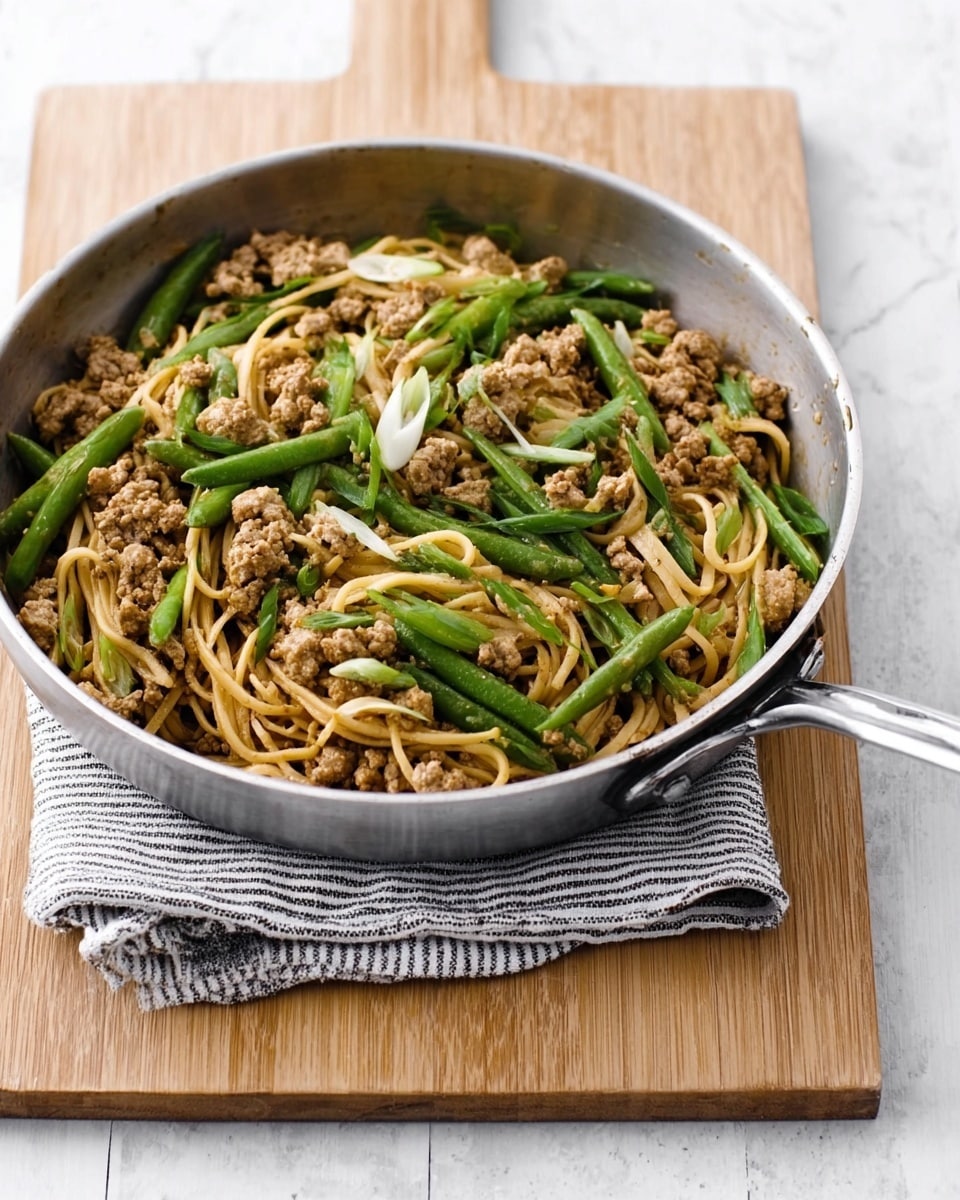 The image shows a round metal frying pan filled with a stir-fry dish resting on a folded striped gray cloth, which is placed on a light wooden cutting board. Inside the pan, there are light brown cooked ground meat pieces mixed with tan-colored noodles at the base. Scattered on top are bright green sliced vegetables like beans and scallions, adding a fresh look to the dish. The background is a white marbled texture, giving a clean and simple setting. photo taken with an iphone --ar 4:5 --v 7
