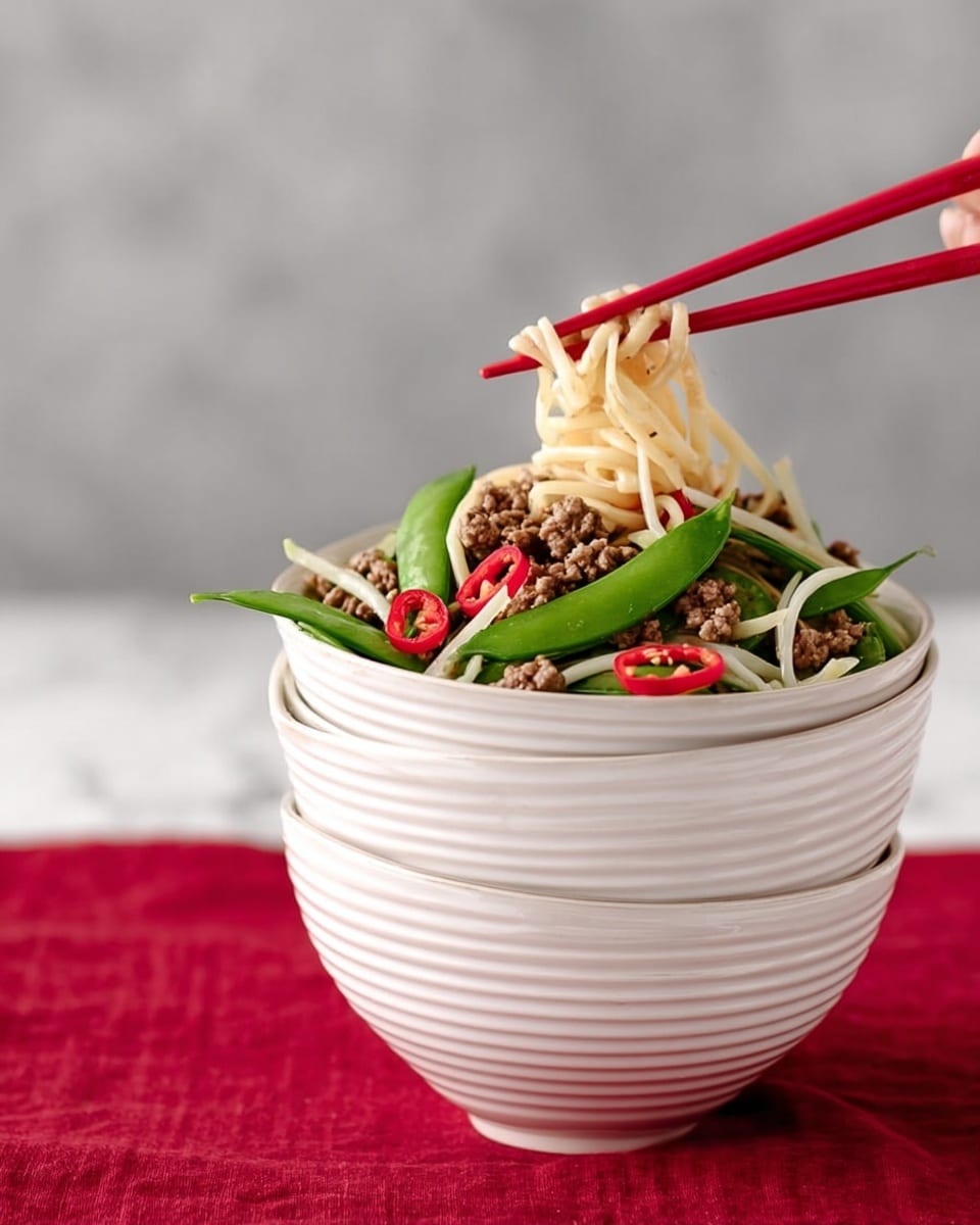 A stack of four white bowls with a ribbed texture is placed on a white marbled surface covered by a red cloth. The top bowl is filled with a mix of food layers: flat beige noodles on the bottom, topped with bright green snap peas, thinly sliced white onions, small pieces of cooked brown ground meat, and thin red chili slices scattered on top. Two red chopsticks are held by a woman's hand and are picking up some noodles from the top bowl. The background is a soft gray color. Photo taken with an iphone --ar 4:5 --v 7