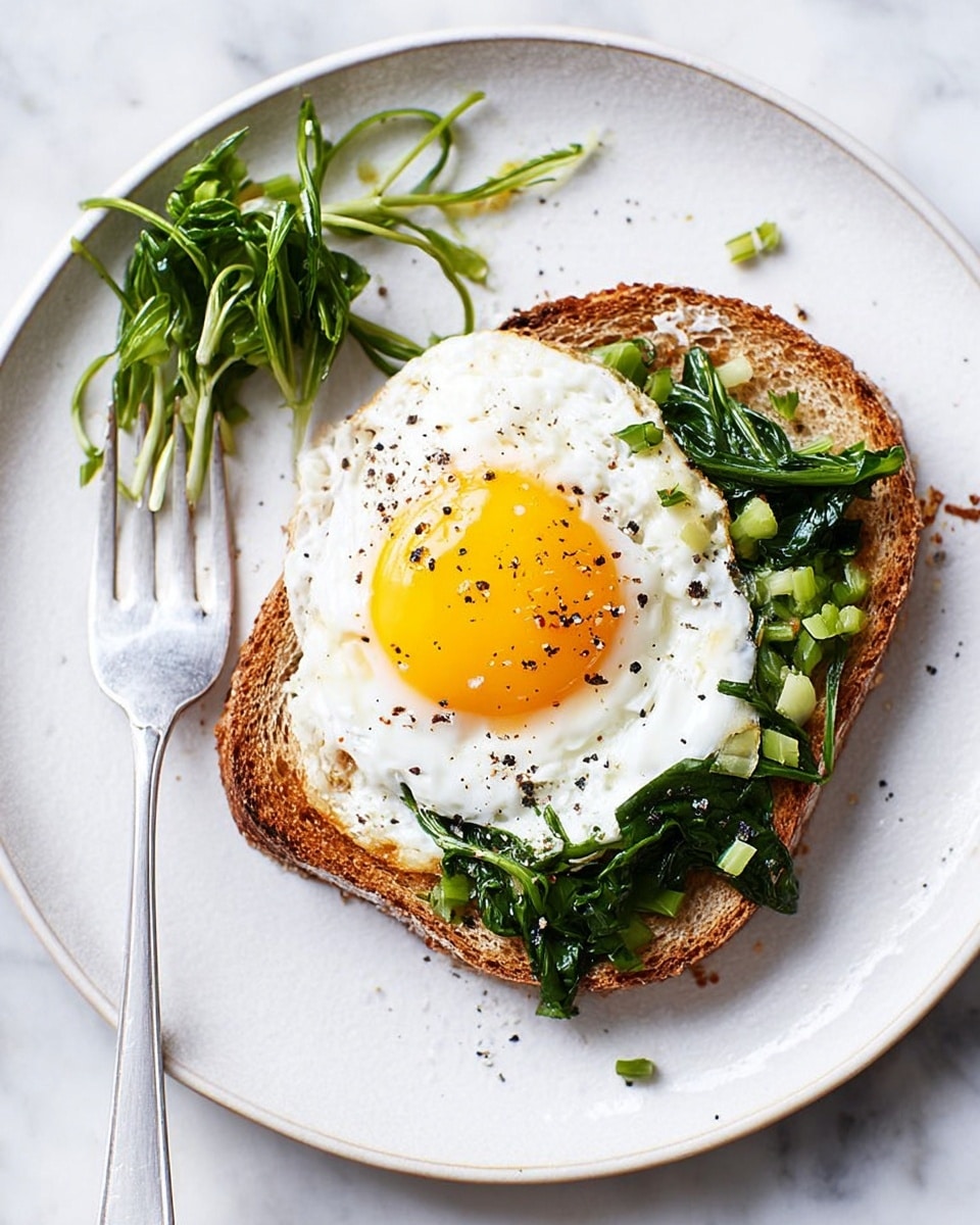 A white round plate on a white marbled surface holds a single slice of toasted brown bread as the bottom layer. On top of the toast is a sunny-side-up fried egg with a bright yellow yolk in the center and white cooked edges. Around the lower right side of the egg, there are chopped green leafy vegetables spread partially on the egg and the toast. Some black pepper is sprinkled lightly over the egg and the plate. A white fork with a small bunch of the same green vegetables is placed on the upper left side of the plate. Photo taken with an iphone --ar 4:5 --v 7