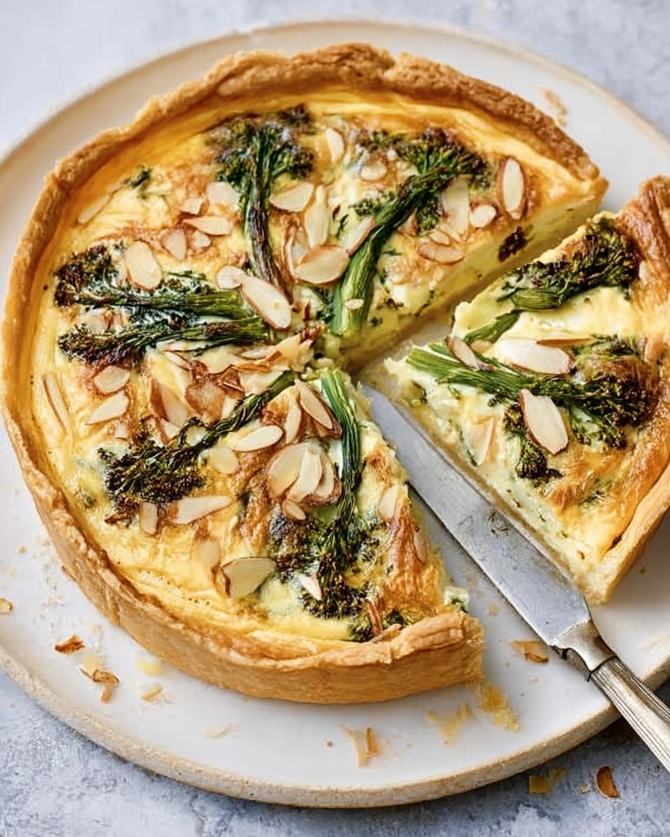 A golden brown round tart with a thick, flaky crust on a white plate. The tart has a creamy pale yellow filling topped with green broccoli or broccolini pieces and scattered almond slices that add texture. A slice is cut out, revealing the smooth filling beneath the vegetables. A silver knife lies beside the plate on a white marbled surface. photo taken with an iphone --ar 4:5 --v 7