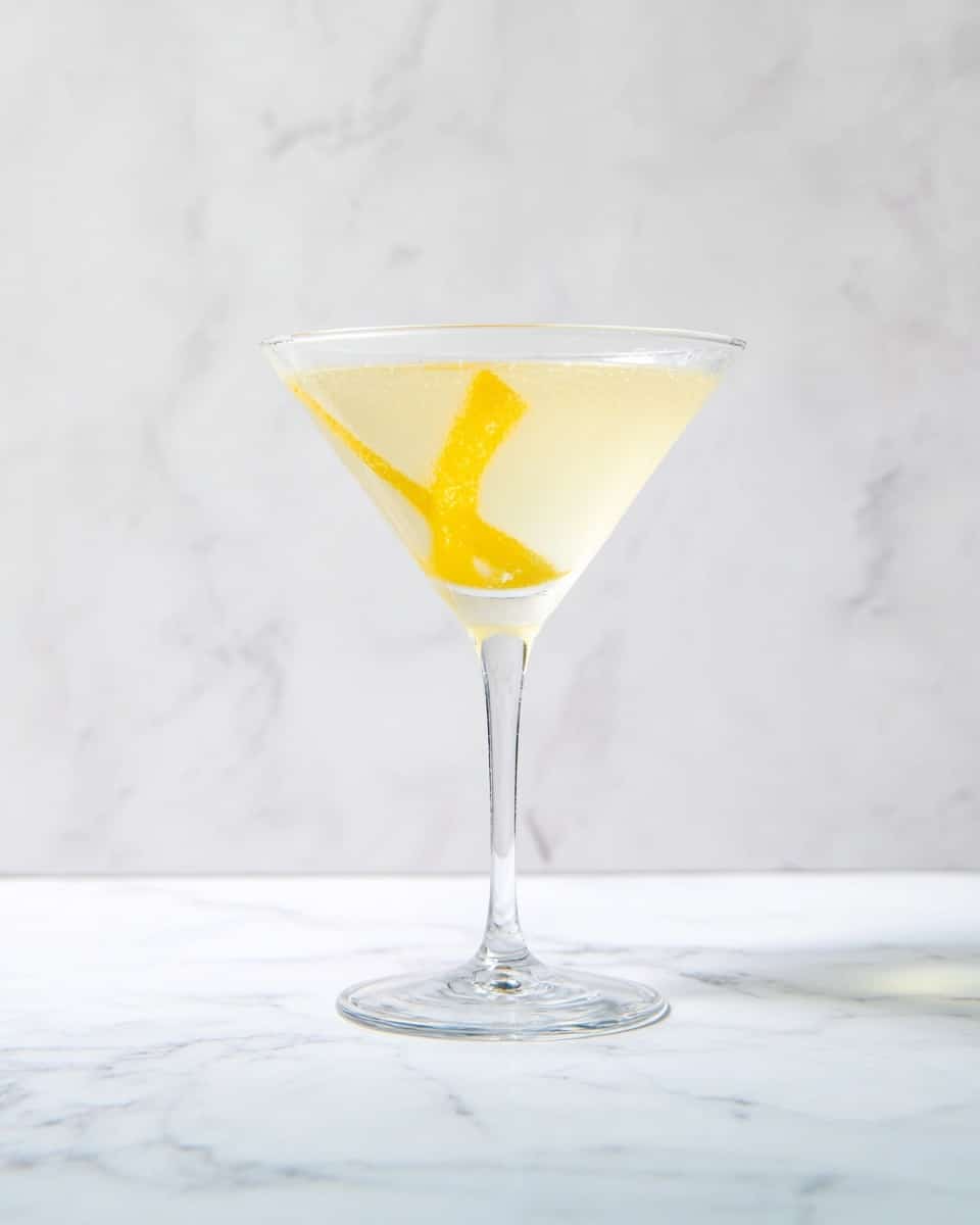 A clear martini glass sits centered on a white marbled surface, filled with a pale yellow cocktail that has a slightly frosted texture. Inside the drink, a thin twist of lemon peel curls gently, adding a bright yellow accent that contrasts with the smooth, translucent liquid. The glass has a long, slender stem and a wide, conical bowl that tapers to a delicate rim. The background is plain white with no distractions, highlighting the elegance and simplicity of the drink. Photo taken with an iphone --ar 4:5 --v 7