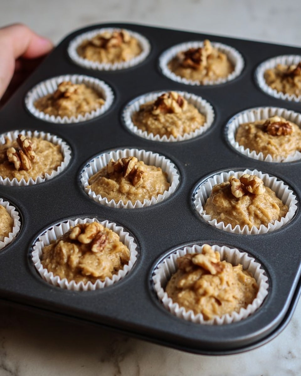 A dark gray muffin tin holds twelve white paper liners filled with light brown, chunky batter. Each scoop of batter is uneven with some texture and topped with one or two walnut halves. The muffin tin rests on a smooth white marbled surface, and the focus is sharp on the nearest cups, with the rest softly blurred in the background. Photo taken with an iphone --ar 4:5 --v 7