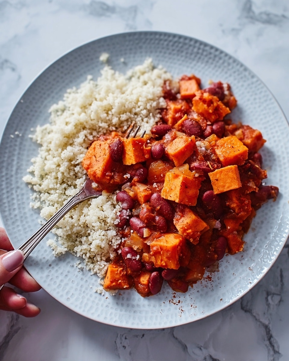 A white plate holds a bed of light, fluffy couscous that looks soft and grainy. On top is a thick layer of chunky chili with pieces of orange sweet potato, reddish-brown beans, and a rich red sauce, giving a hearty and warming feel. A silver fork rests partly in the couscous and chili, angled towards the bottom left of the plate. The plate is on a white marbled surface with soft natural light highlighting the textures and colors. photo taken with an iphone --ar 4:5 --v 7