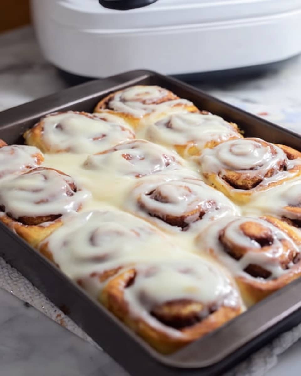 A rectangular pan filled with a dozen cinnamon rolls, each roll showing visible layers of golden-brown dough spiraled with a darker cinnamon filling. The rolls are covered with a thick layer of creamy white icing that drapes smoothly over the top and slightly between the swirls, giving a glossy and slightly melted look. The pan sits on a white marbled surface with a blurred white appliance in the background. photo taken with an iphone --ar 4:5 --v 7