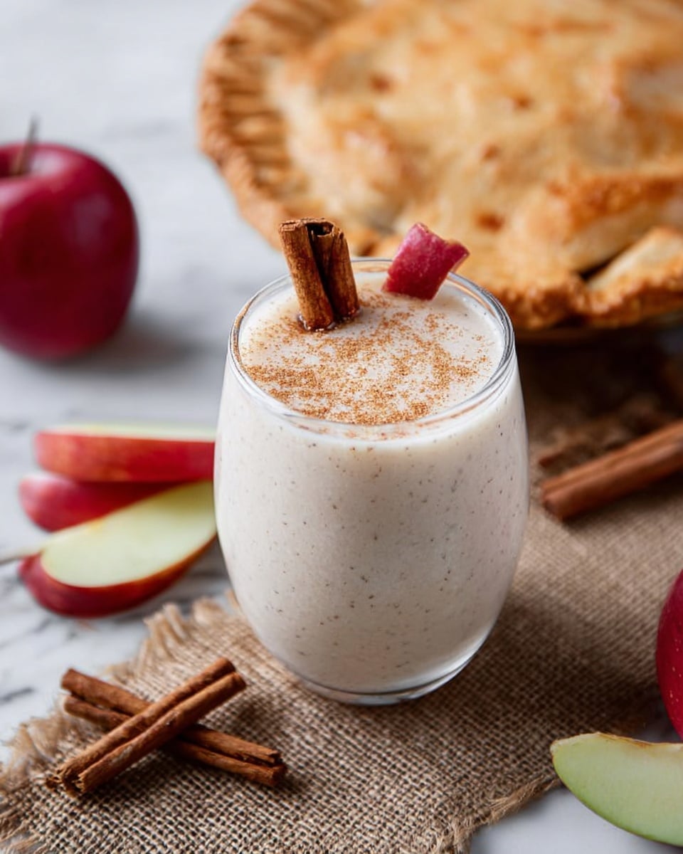 The image shows a clear glass filled with a thick, creamy beige drink, topped with a dusting of cinnamon powder. A cinnamon stick and a small red apple slice rest on the rim of the glass. Next to the glass on a textured burlap cloth, there is a golden-brown baked pie with a slightly cracked top and cinnamon sticks scattered around it. Additional red and green apple slices are placed casually near the pie. The scene is set on a white marbled surface. Photo taken with an iphone --ar 4:5 --v 7