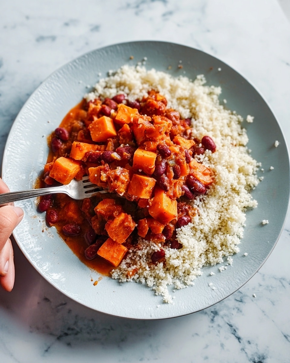 The image shows a white plate with two main layers of food. The bottom layer is a fluffy, white, grain-like texture spread across the plate’s base. On top is a thick stew made up of bright orange cube-shaped pieces, likely sweet potatoes, mixed with small kidney beans and other small beans in a rich, deep red sauce. A fork rests on the plate, partially inserted into the stew, with a woman's hand holding it. The background is a white marbled surface. Photo taken with an iphone --ar 4:5 --v 7