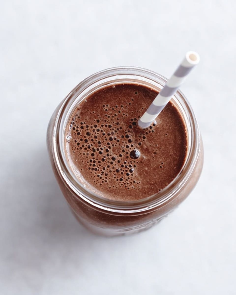 The image shows a close-up, top view of a glass jar filled with a chocolate-colored smoothie. The smoothie is thick and has tiny bubbles on its surface. A white straw with light gray stripes is placed diagonally inside the jar. The jar sits on a white marbled surface, giving it a clean and bright background. photo taken with an iphone --ar 4:5 --v 7