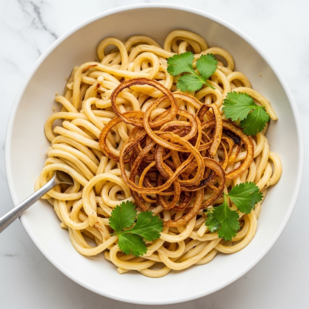 The image shows a white bowl filled with a single layer of thick, creamy white noodles. On top of the noodles, there is a scattered layer of fresh green chopped herbs, likely cilantro, adding a bright pop of color. Sitting neatly in the center, there is a small pile of golden-brown, thin fried strips, likely fried onions or similar, with a slightly crispy texture. A metal fork is partially visible, resting inside the bowl. The background is a white marbled texture. photo taken with an iphone --ar 4:5 --v 7