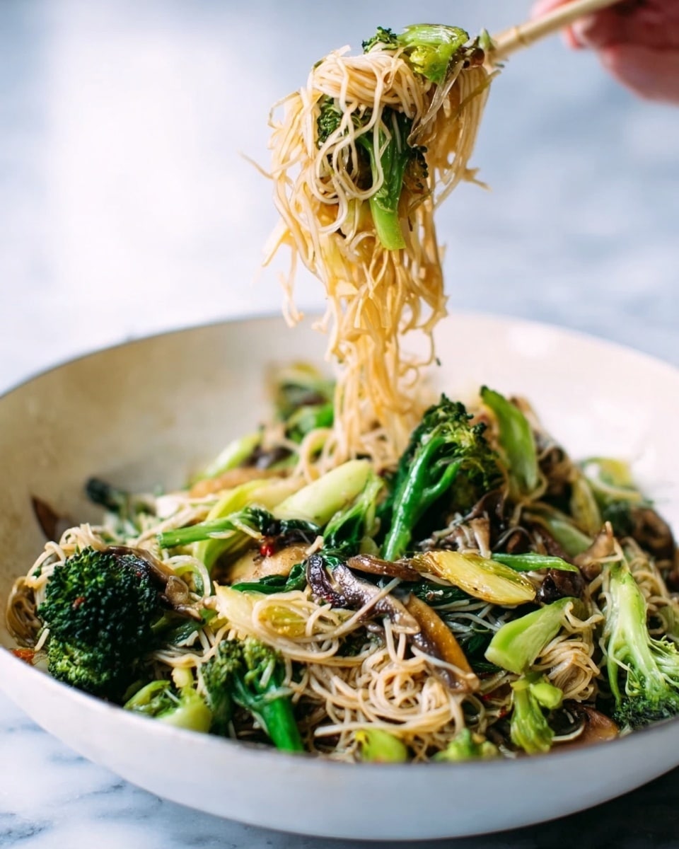 The image shows a close-up of a large white bowl filled with a noodle dish that has thin, light-colored noodles mixed with dark green broccoli florets, small pieces of light green vegetables, and brown mushrooms. A woman's hand is using wooden tongs to lift the noodles, which hang loosely and show the mix of vegetables underneath. The white marbled surface can be seen blurred in the background. photo taken with an iphone --ar 4:5 --v 7
