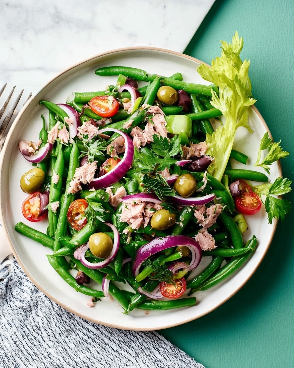 A large white plate holds a colorful salad with several layers visible: at the base are long green beans, mixed with bright green olives and small reddish-brown nuts scattered throughout. Thin, curved slices of purple-red onion are spread evenly over the salad along with light green celery leaves and fresh flat green parsley leaves on top, adding different textures and colors. Small chunks of light pink tuna are also mixed in among the vegetables, giving the salad a varied look. The plate sits on a white marbled surface. photo taken with an iphone --ar 4:5 --v 7
