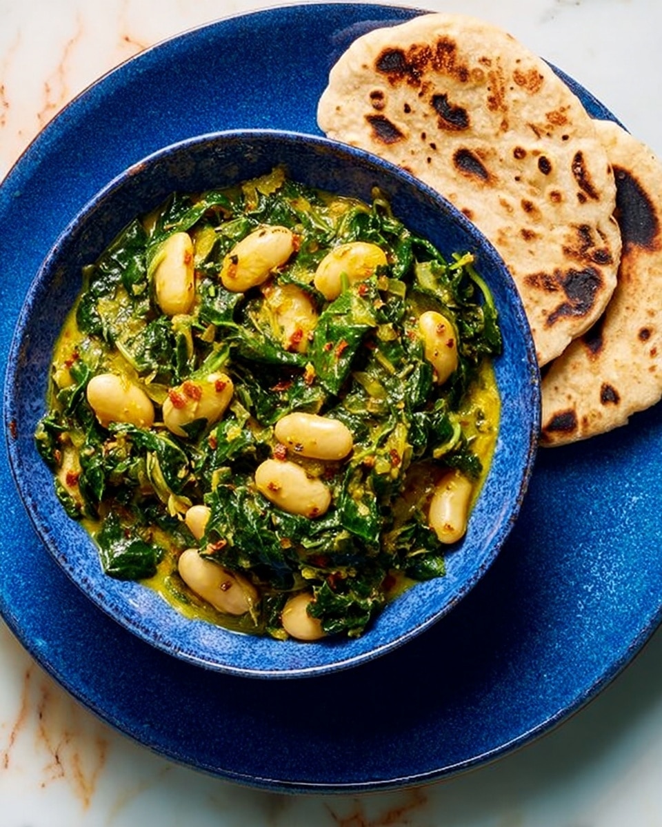 A blue bowl filled with a mix of cooked leafy greens and large white beans, coated in a yellowish sauce with small bits of spices or herbs visible throughout, placed on a larger blue plate. Next to the bowl are two pieces of flatbread with char marks and soft texture, resting directly on a white marbled surface. The colors are mainly green from the leaves, white from the beans, and yellow from the sauce, with blue plates creating a strong contrast. photo taken with an iphone --ar 4:5 --v 7